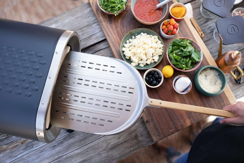 A close-up, top-down view of a perforated metal pizza peel sliding under a perfectly baked Neapolitan-style pizza to remove it from a hot home pizza oven.
